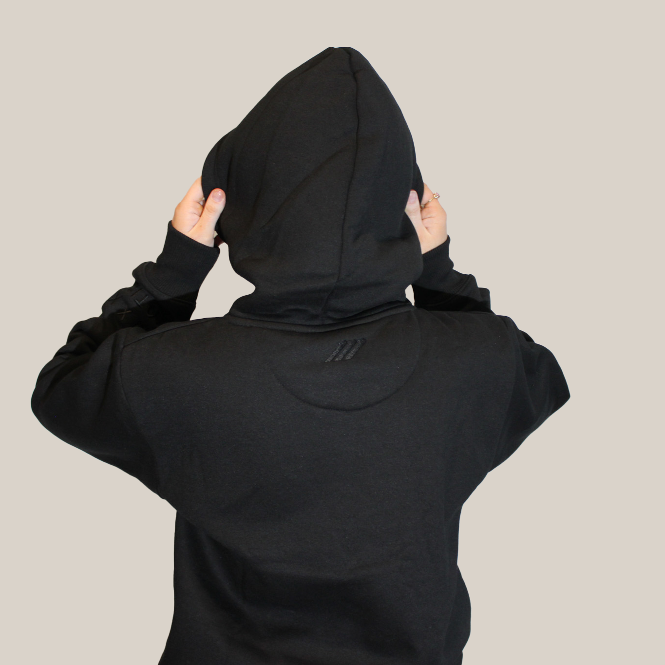 Person wearing a black hoodie with embroidery Motion logo on the back neckline with the hood up on a beige background